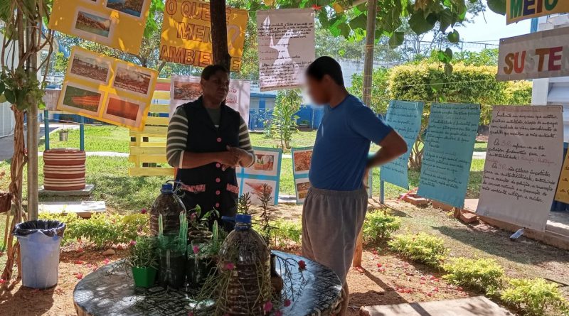 Fotografia retrata educadora e adolescente conversando, em meio a um gasebo coberto por folhas de plantas. No ambiente, que fica na área externa, estão afixados cartazes sobre o tema e há plantas na mesa de centro, que é uma bobina de fios reciclada. O ambiente é sombreado e repleto de verde