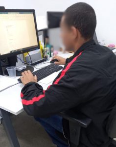Fotografia retrata adolescente de costas, mexendo em um computador. O adolescente usa uma blusa de frio preta com uma listra vermelha ao longo do braço. 