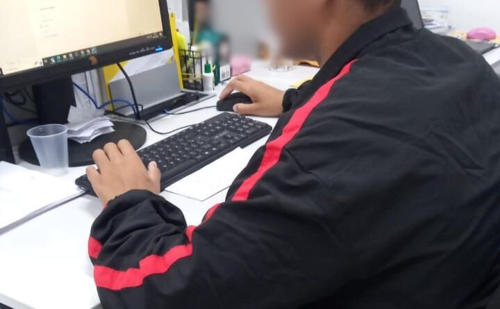 Fotografia retrata adolescente de costas, mexendo em um computador. O adolescente usa uma blusa de frio preta com uma listra vermelha ao longo do braço.